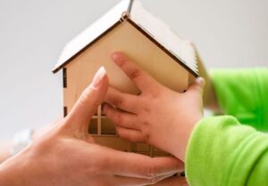 Close-up of mother and child holding a small wooden model house, symbolizing family and home.