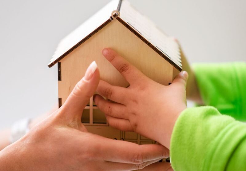 Close-up of mother and child holding a small wooden model house, symbolizing family and home.